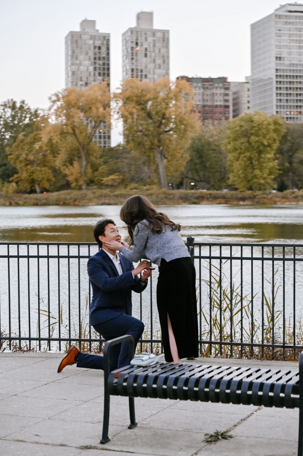 People interact on park benches along a waterfront with autumn trees and city buildings in view.