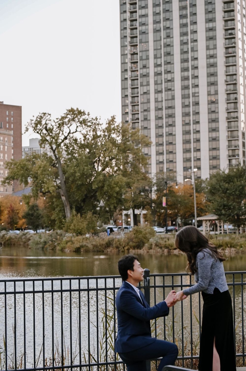 A romantic proposal scene unfolds by a lake with high-rise buildings and fall foliage in the background.