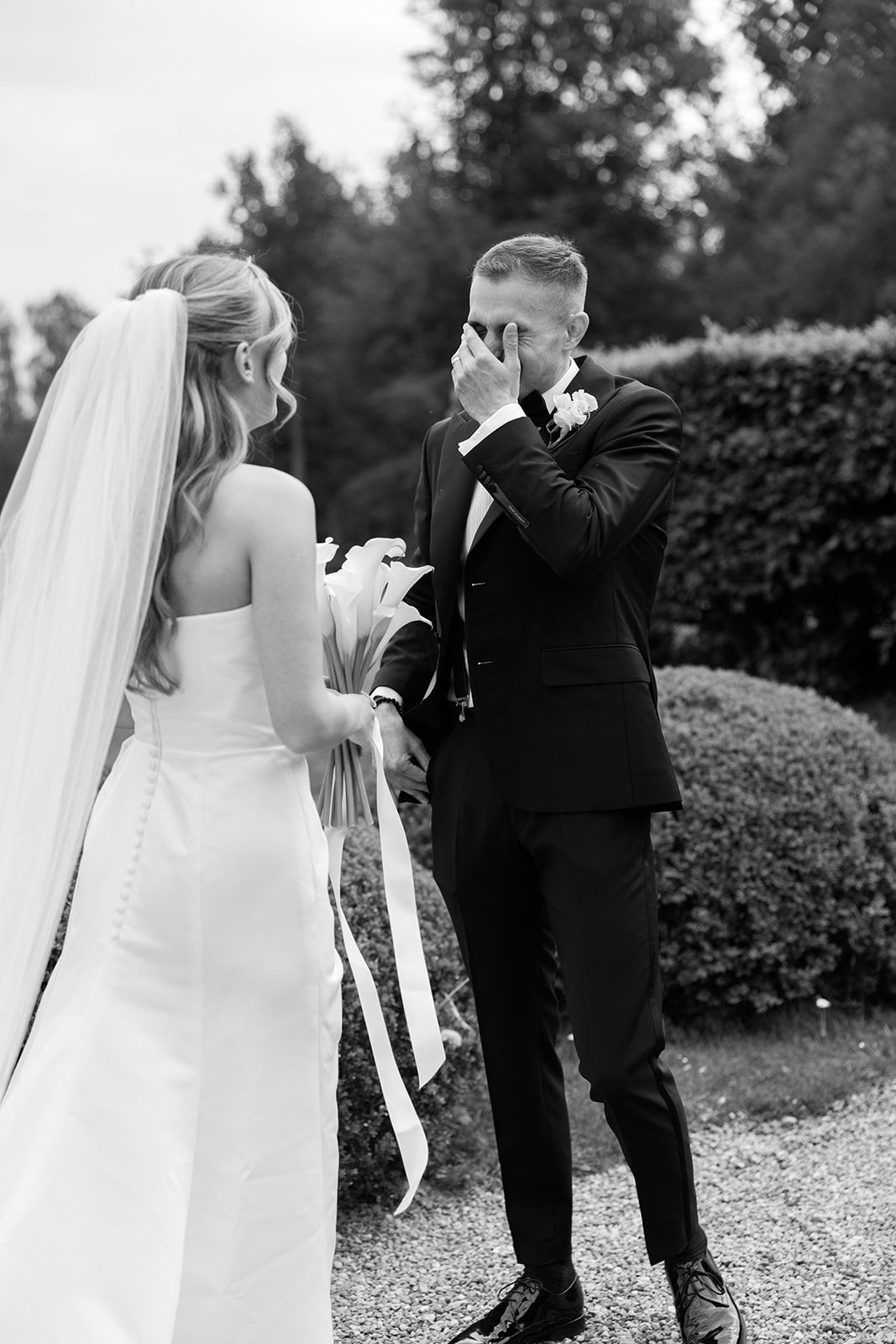 A bride playfully kicks the groom from behind during an outdoor wedding photoshoot on a gravel path.