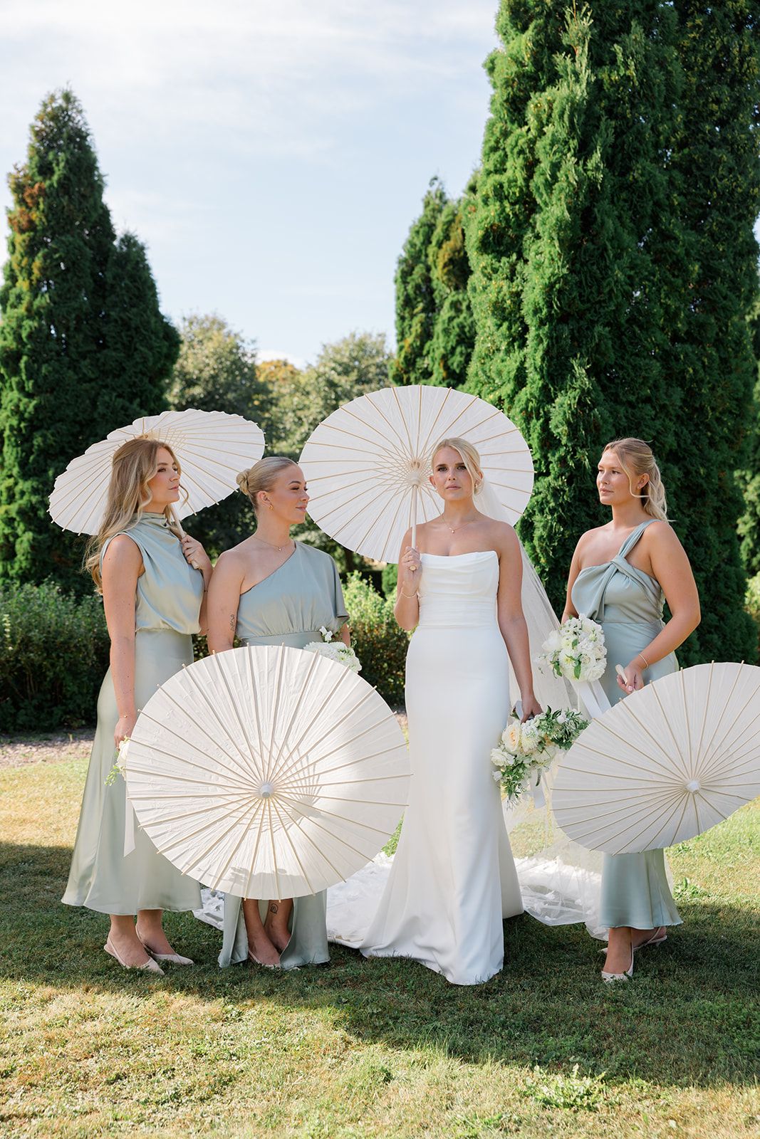 A group in light blue dresses poses outdoors with white paper parasols against a backdrop of tall green cypress trees.