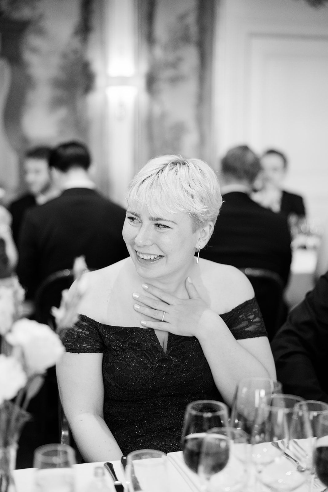 A person in a black dress smiles and laughs during a formal dinner event in black and white photography.