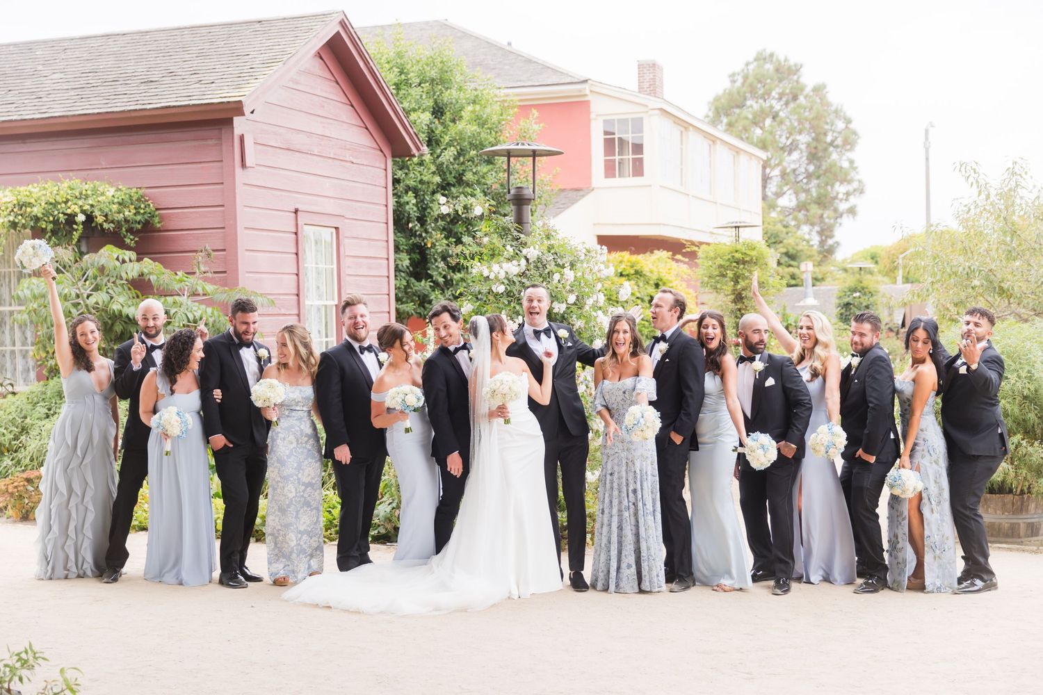 Wedding party in pink bridesmaid dresses and black tuxedos pose together on a sunny lawn under shady trees.