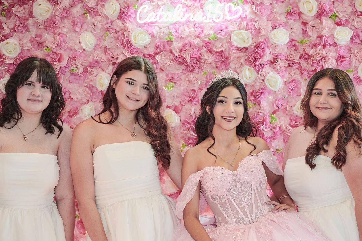 A pink floral wall serves as a backdrop for people wearing white formal dresses posing together at an event.