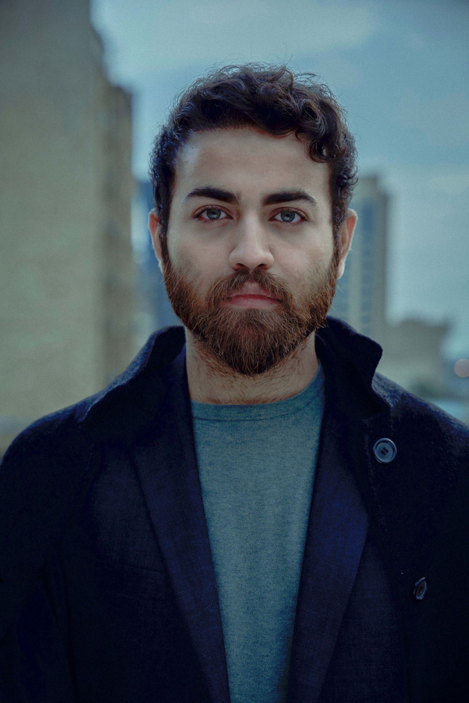 A person with a dark beard wearing a black coat and gray sweater gazes at the camera in a series of similar portraits.