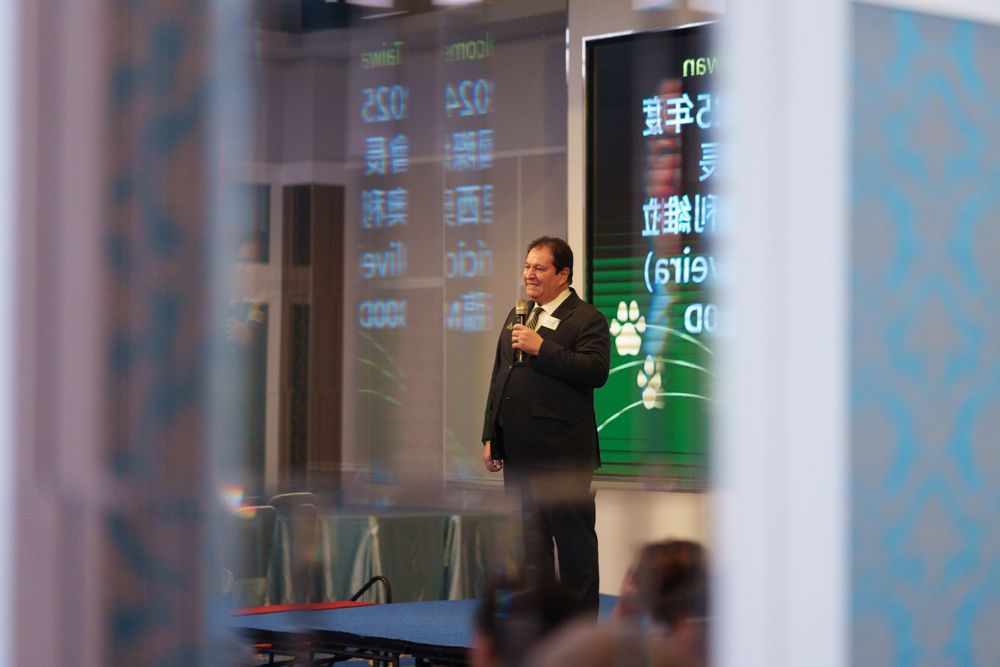 A person in a suit stands in front of a glass wall with green digital displays showing financial data.