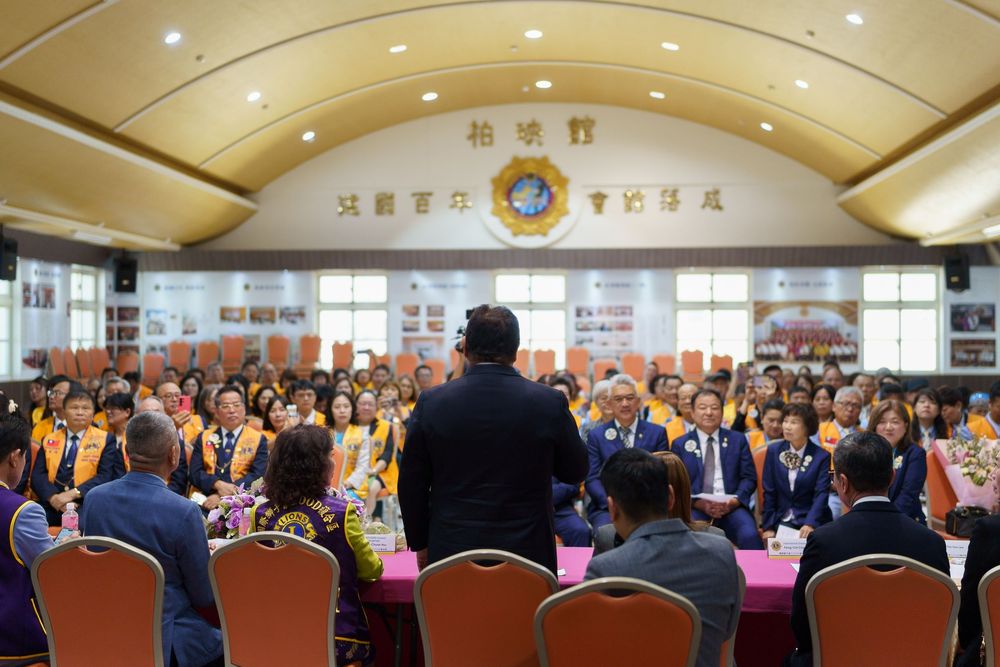 A large conference room filled with attendees during a formal gathering with a decorated ceiling and organizational emblems.