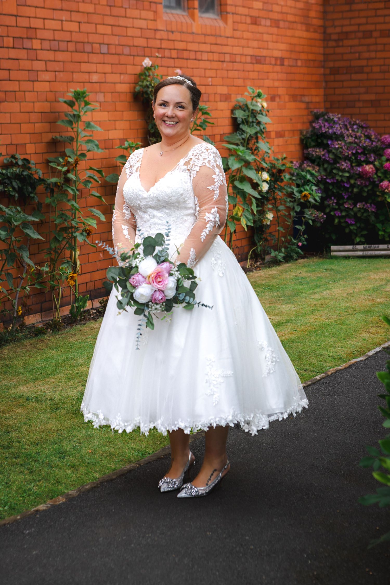 Beautiful bridal portrait in tea-length lace wedding dress with long sleeves holding pink and white bouquet against brick wall.