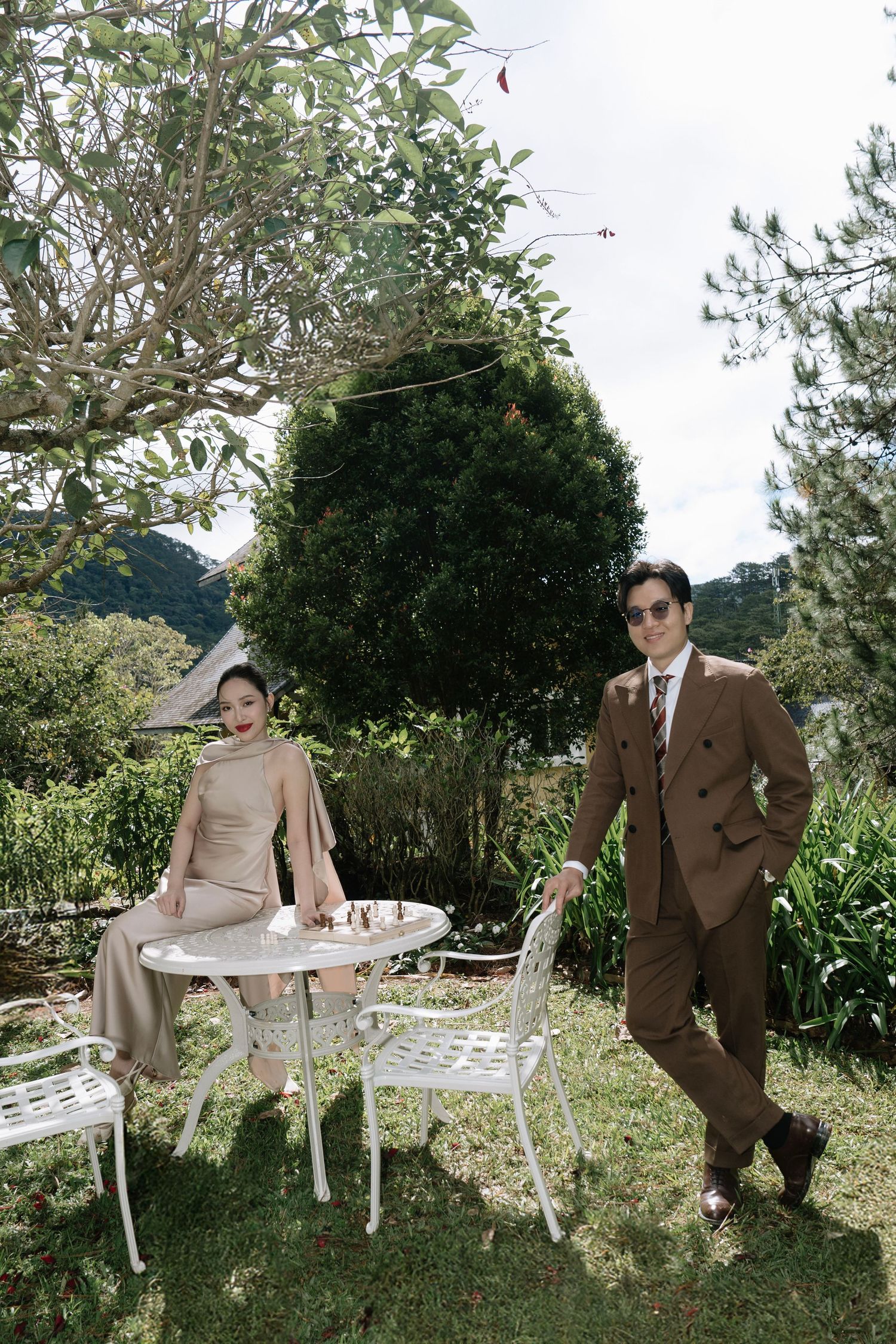 Stylish couple posing beside white garden table with chess set in lush green outdoor setting.