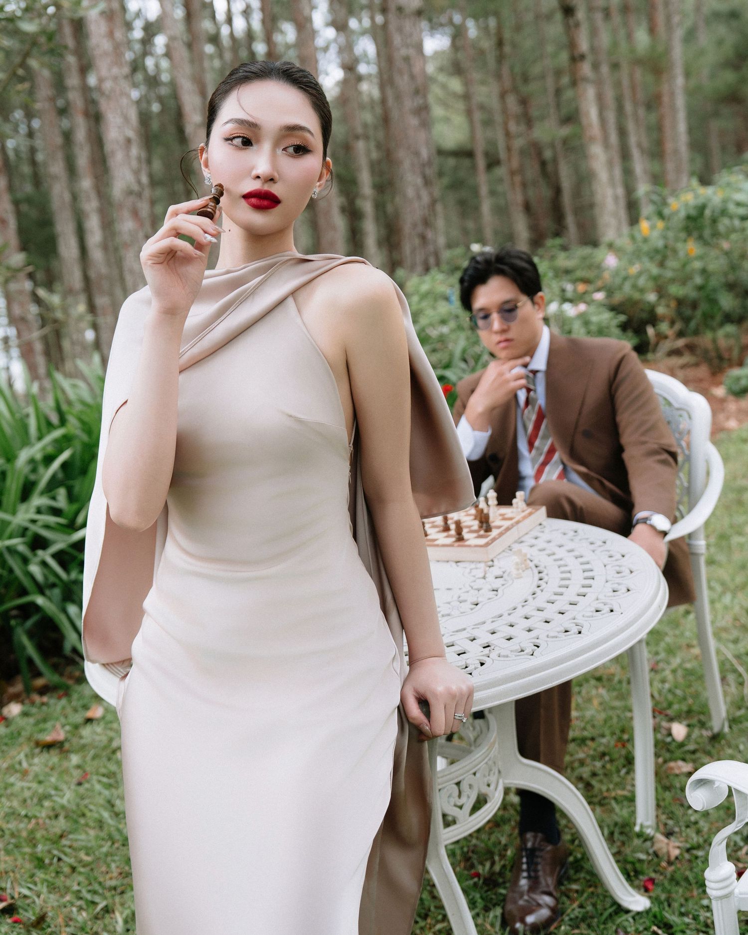 Elegant woman in beige dress holding chess piece with man in brown suit in forest background.