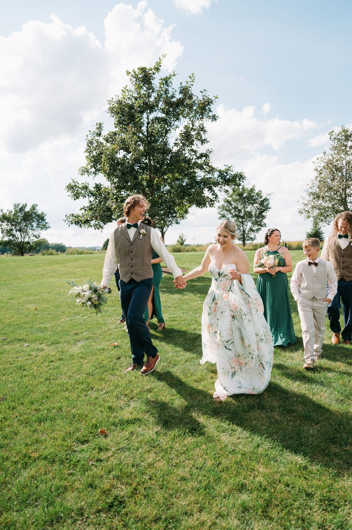 bride and groom holding hands and leading bridal party across green space in Lowe Park Marion Iowa by Iowa midwest wedding photographer