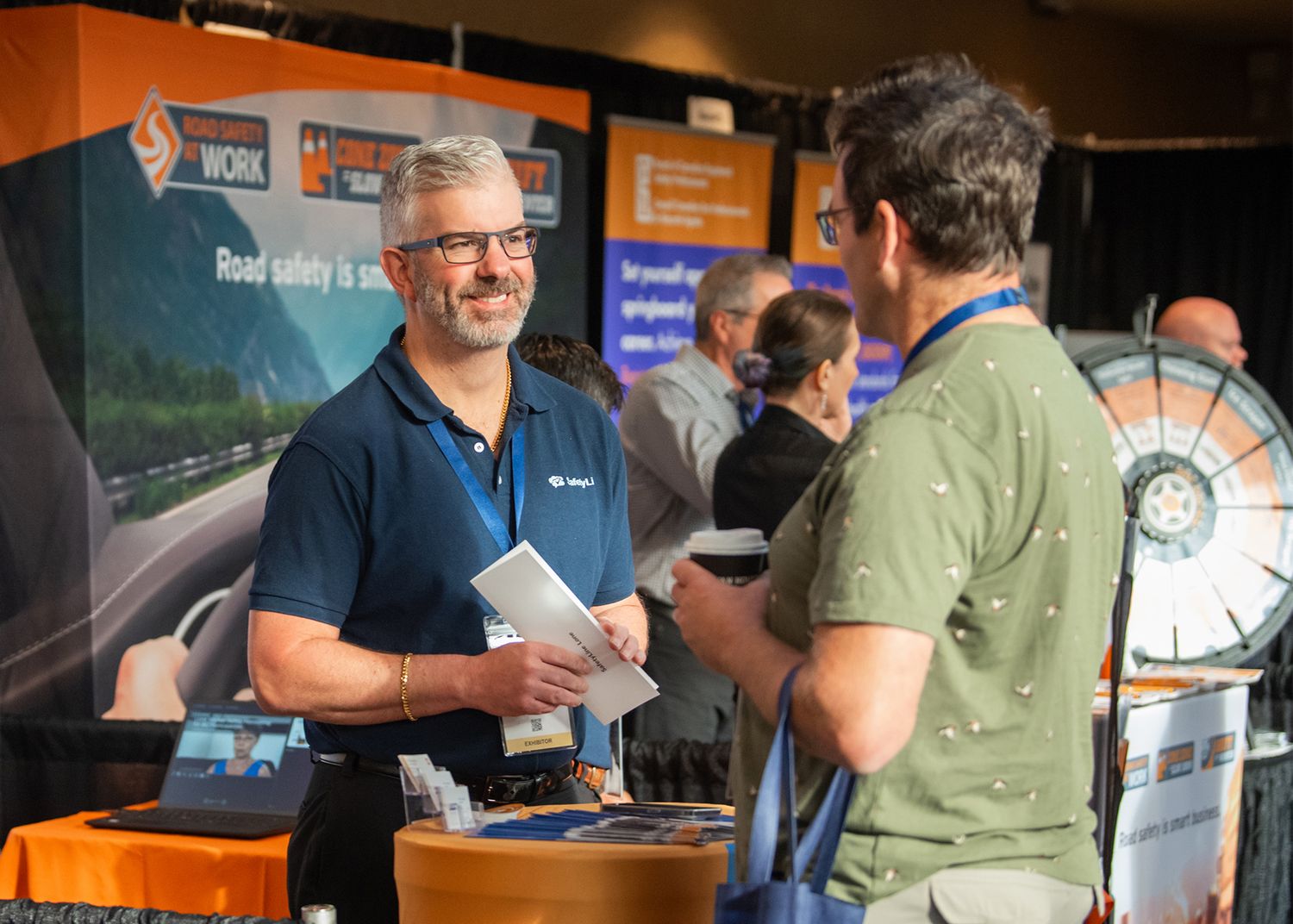 Two men talking at a trade show booth with branded display materials in the background.