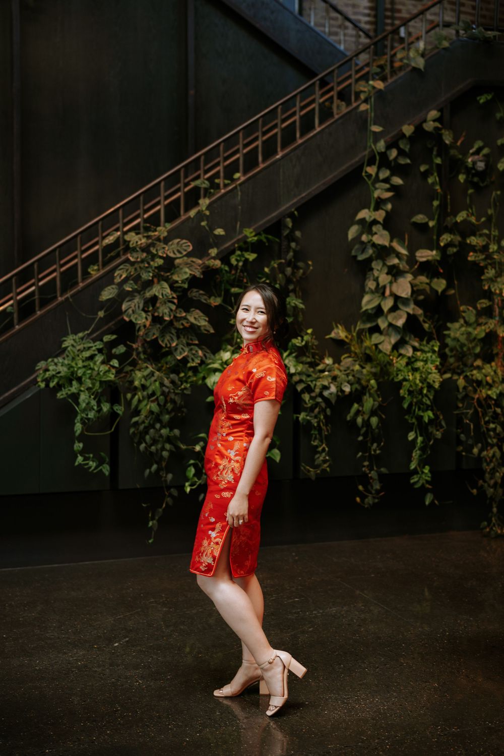 A person wearing a traditional red Chinese qipao dress poses gracefully against a dark background with vines on the wall. Picnic Fishtown
