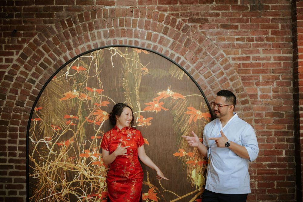 A couple in a red dress and blue shirt poses together against a rustic brick wall with decorative golden branches. Picnic Philadelphia