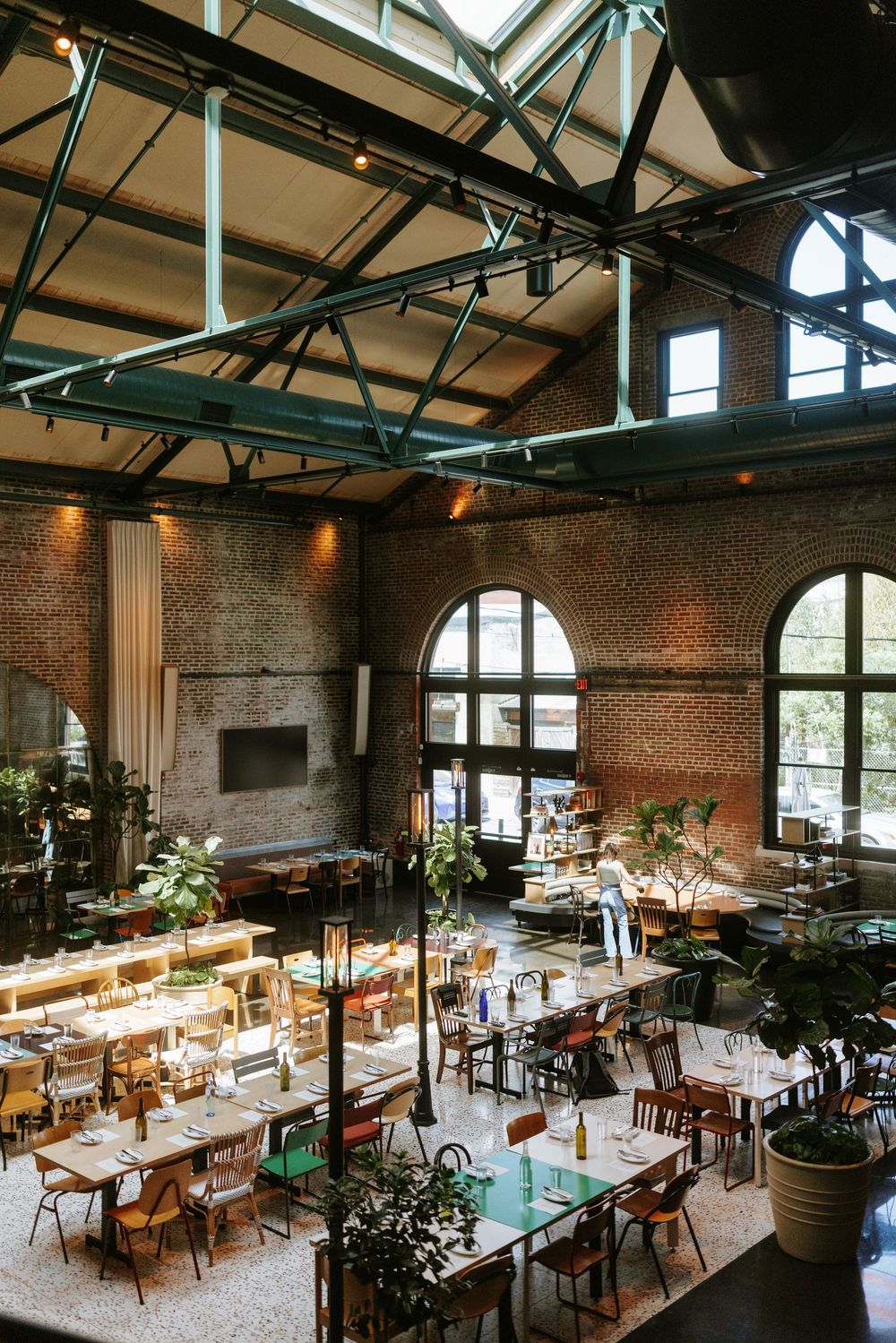 Modern industrial restaurant interior with exposed brick walls high ceilings, and communal wooden dining tables arranged in rows. Picnic