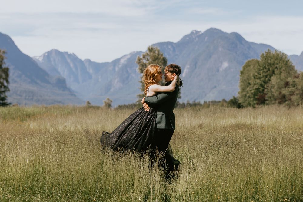 A couple in formal attire embrace in a field with majestic mountain peaks rising in the background.
