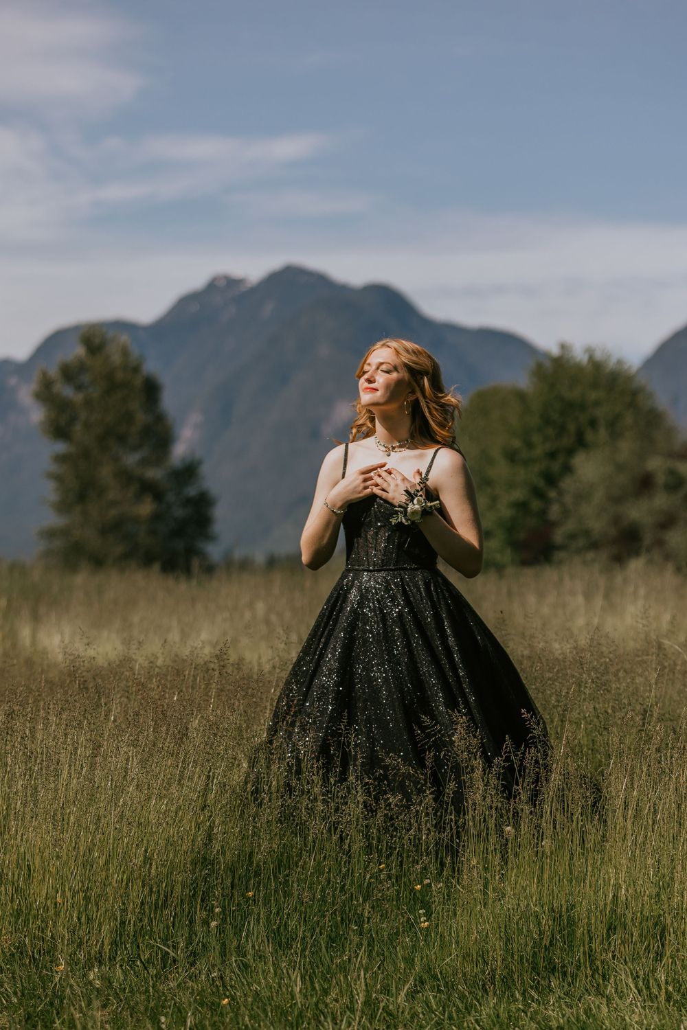 Person in a black flowing dress stands in a tall grass meadow with mountains and cloudy sky in the background. Grad portraits