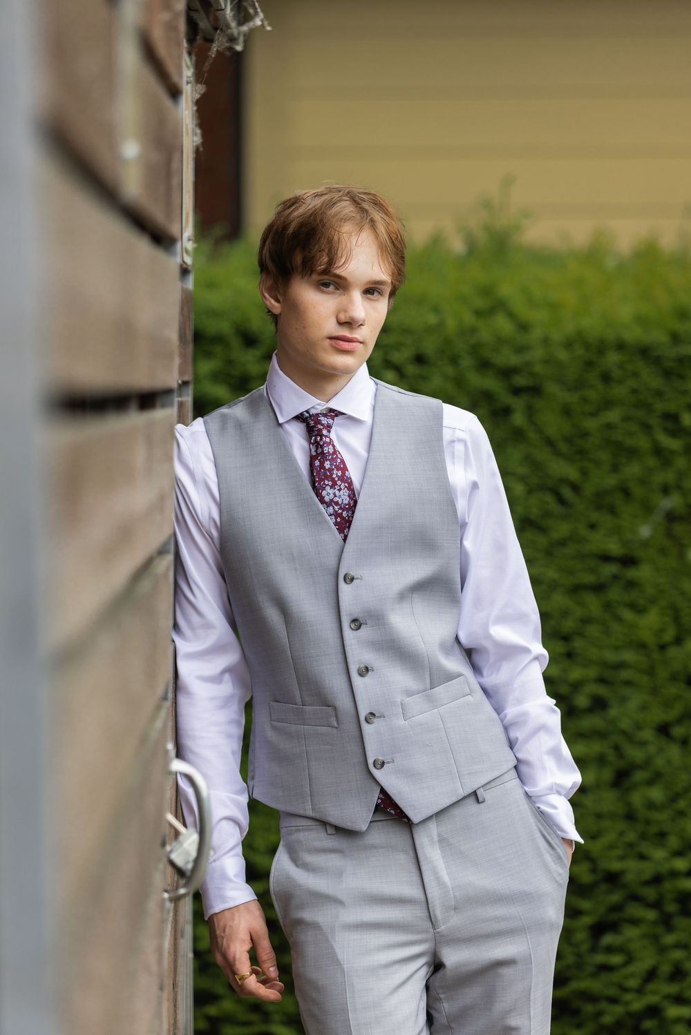 Young man in gray suit vest, white shirt, and floral tie posing outdoors near wooden wall with green hedges.