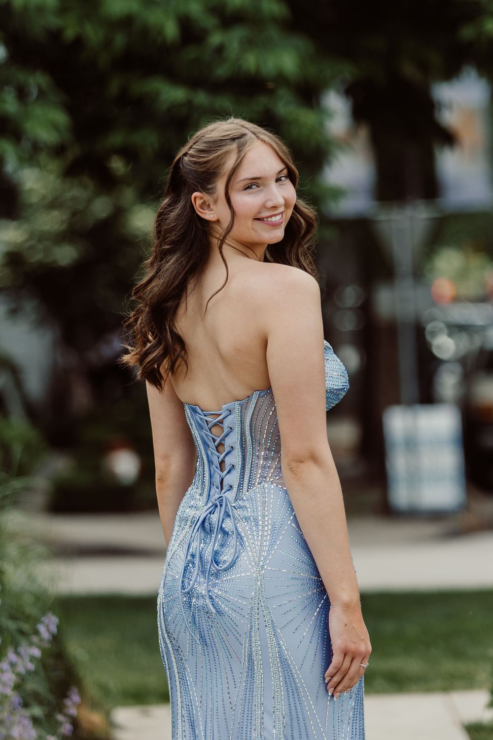 Young woman smiling in a strapless light blue corset-back beaded gown, posing outdoors with lush green trees.