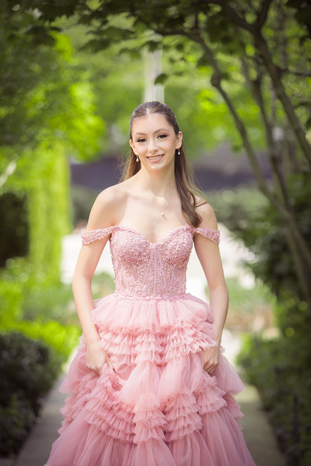 Elegant pink ball gown with ruffles and off-shoulder design photographed in a lush garden setting.