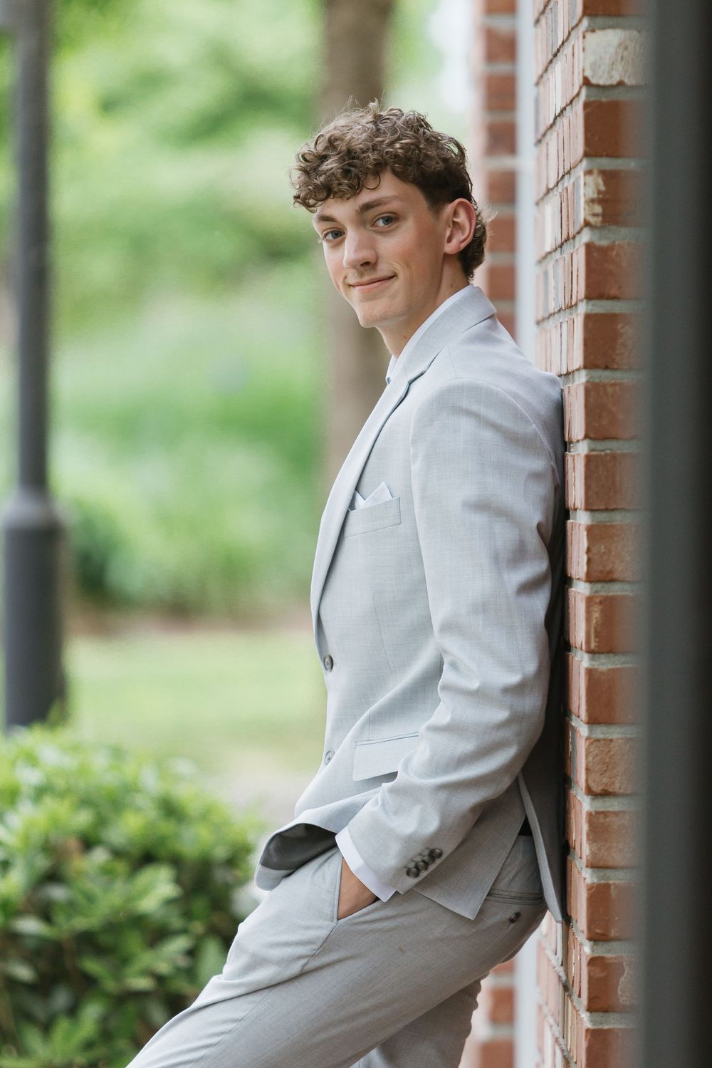 Young man in light gray suit leaning against brick wall outdoors, smiling confidently for a senior portrait photo.