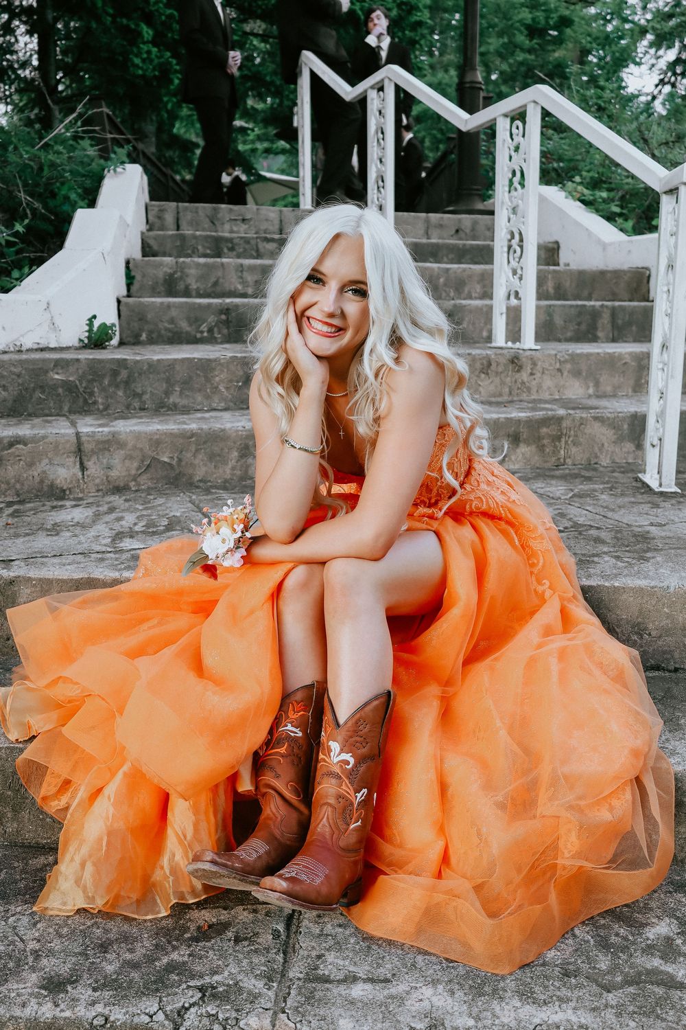 Young woman in orange tulle gown and cowboy boots sits smiling on stone steps holding a floral corsage.