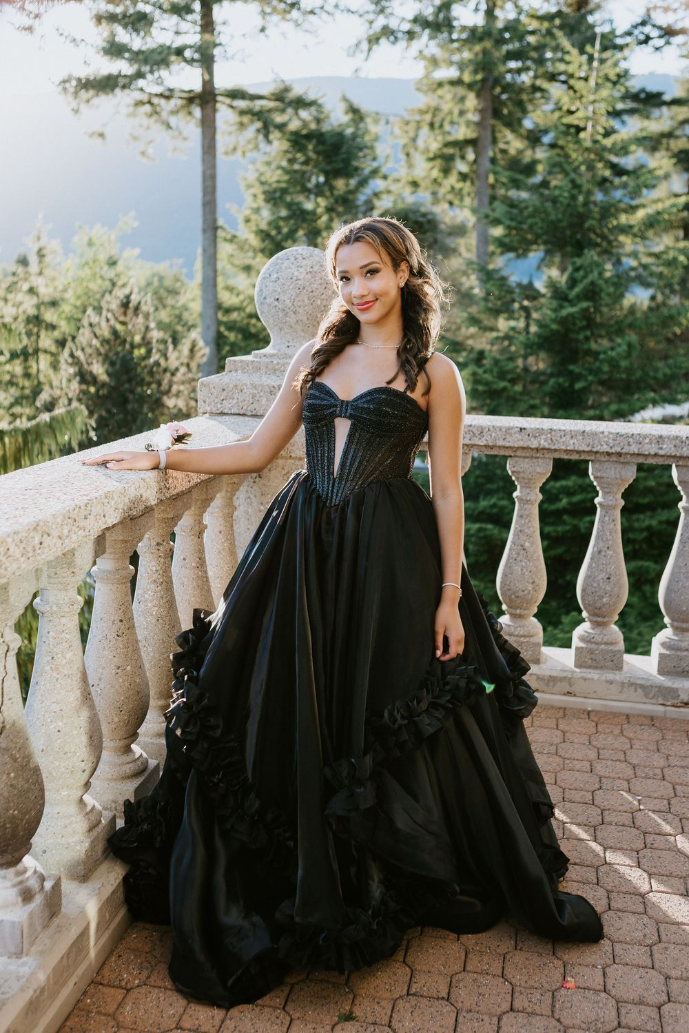 Woman in black strapless ball gown with floral details poses on stone balcony surrounded by lush green trees.