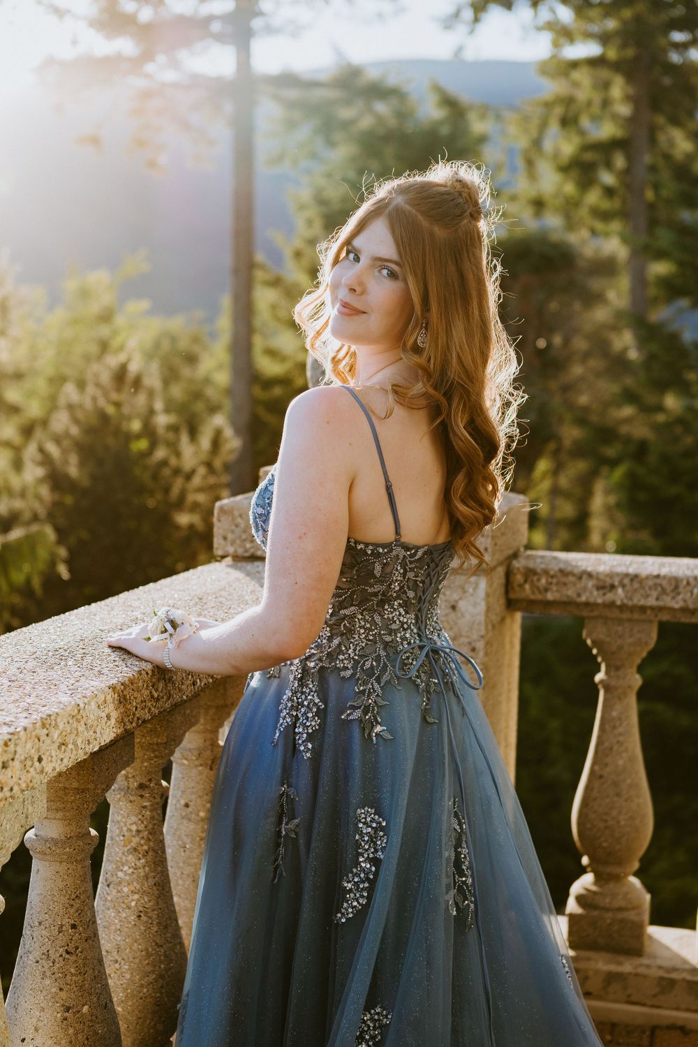 Young woman in elegant blue embroidered gown poses on stone balcony surrounded by lush forest at golden hour.