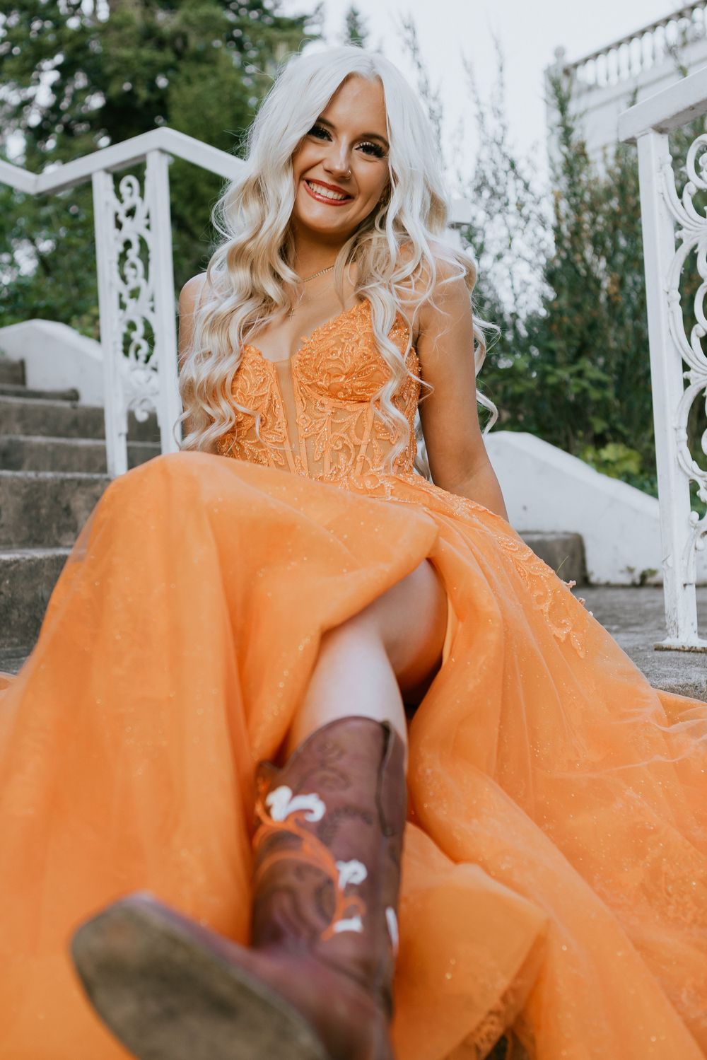 Smiling blonde woman in orange tulle gown and brown cowboy boots sitting on white staircase steps.