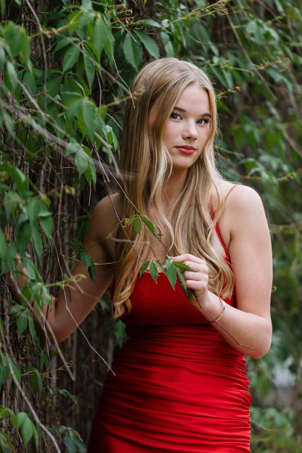 Young blonde woman in a red dress posing gracefully among lush green ivy vines outdoors.