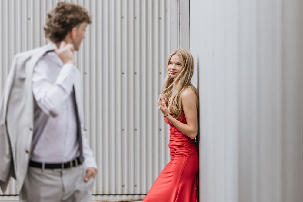 A woman in a red dress leans against a wall, smiling at a man in a gray suit walking past her outdoors.