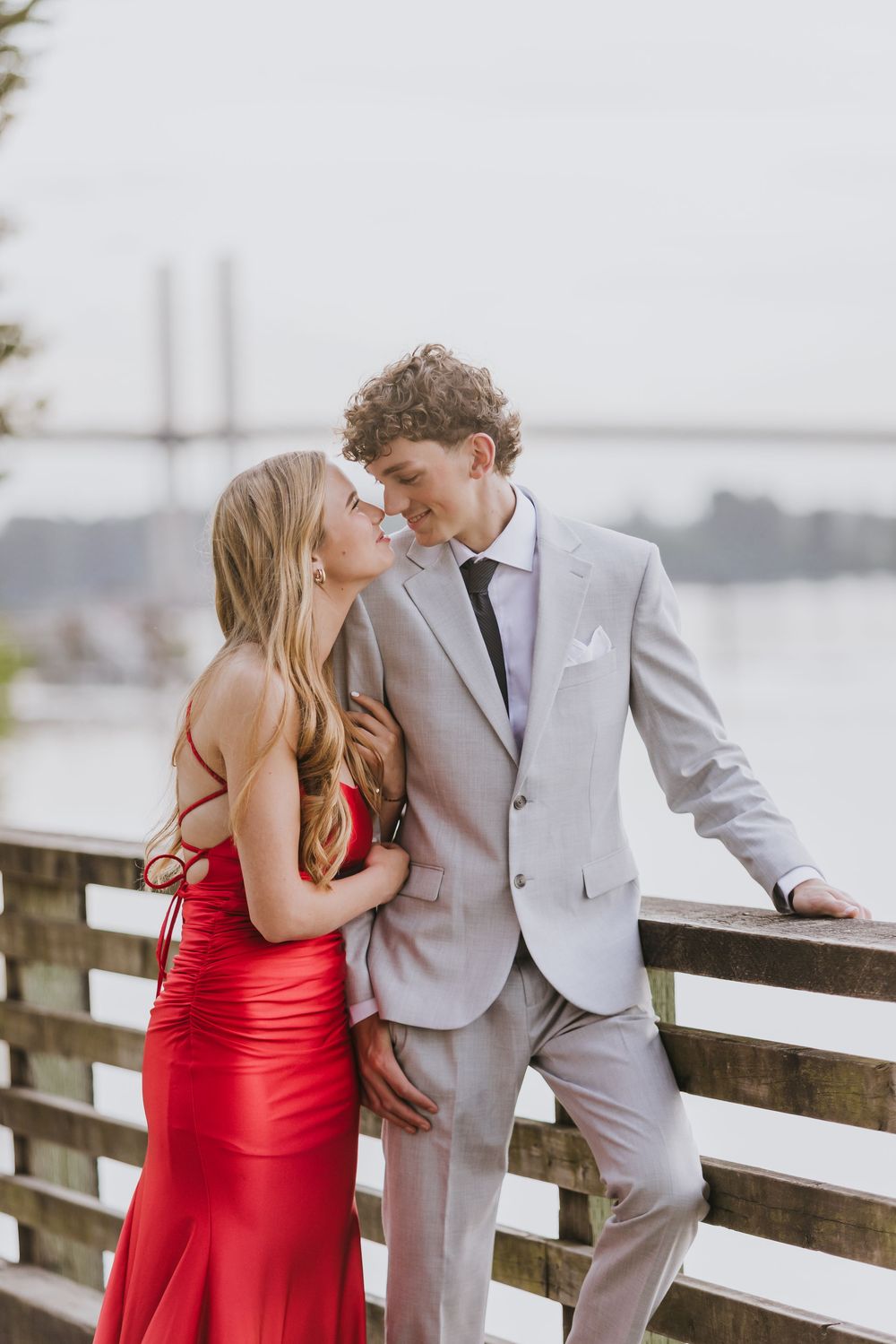 A couple in formal wear, woman in red gown and man in gray suit, share a romantic moment on a wooden bridge.