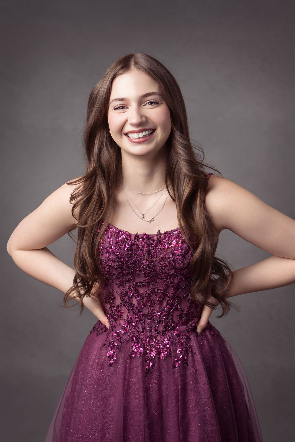 A young person poses in a plum-colored formal dress with sequined bodice and tulle skirt against a gray backdrop.