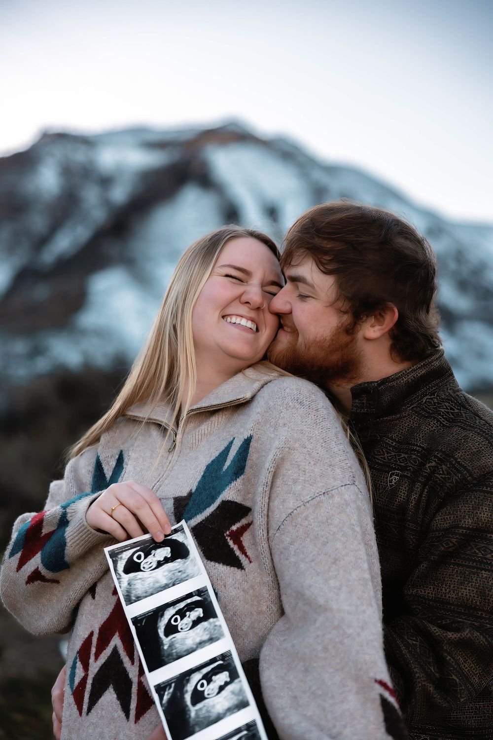 A couple shares an intimate moment while holding ultrasound photos against snowy mountain peaks.