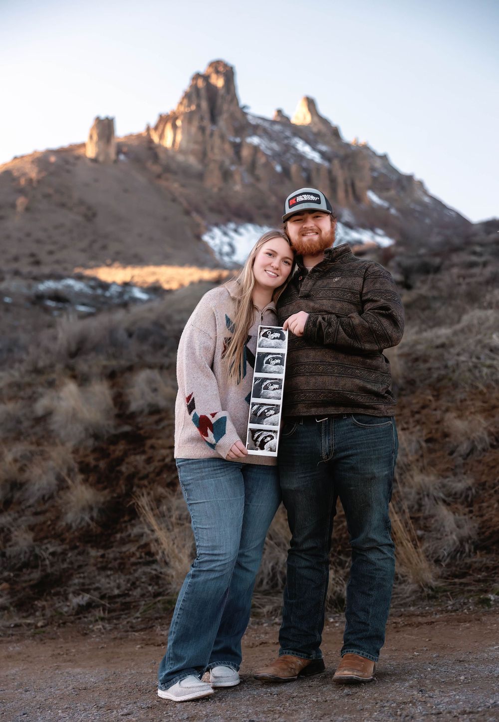 A couple in casual winter wear shares ultrasound photos against a scenic mountain backdrop at sunset.