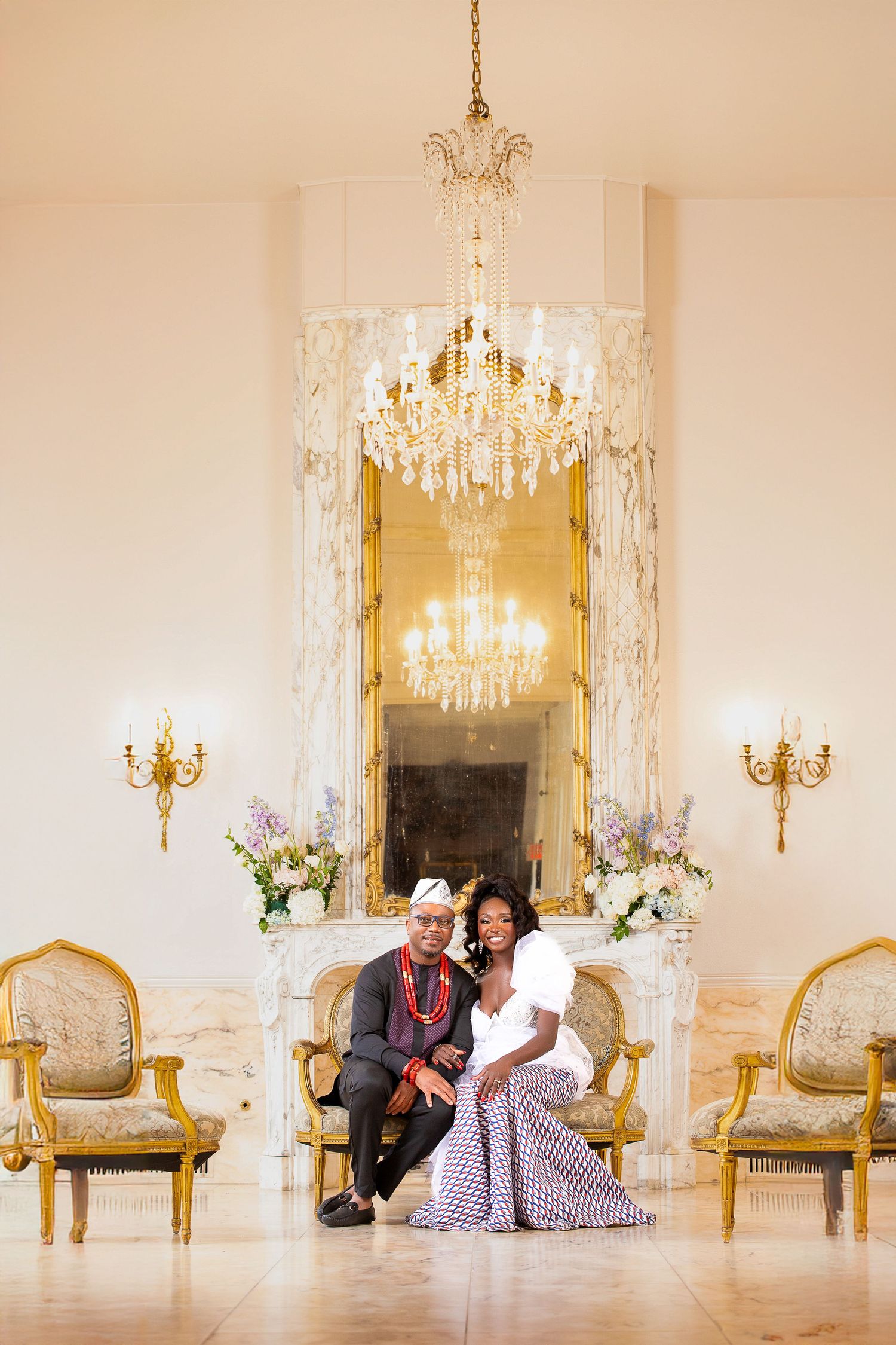 Romantic couple poses together in an elegant white ballroom with crystal chandeliers and ornate gold furniture.