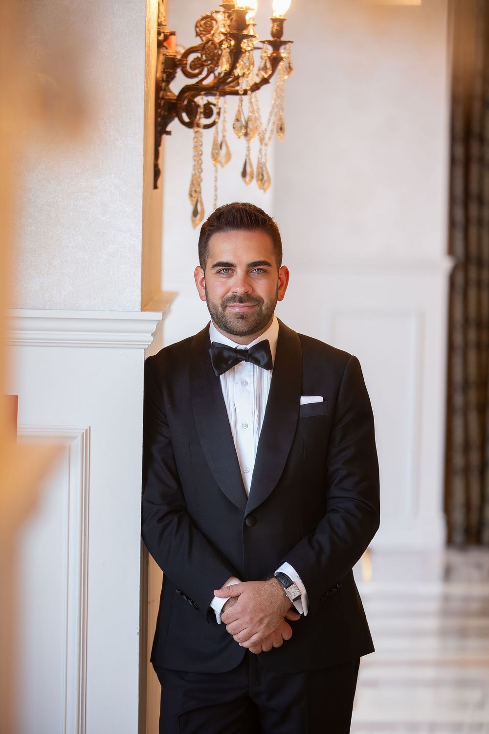 Professional portrait photograph in a formal black tuxedo standing against a light colored wall.