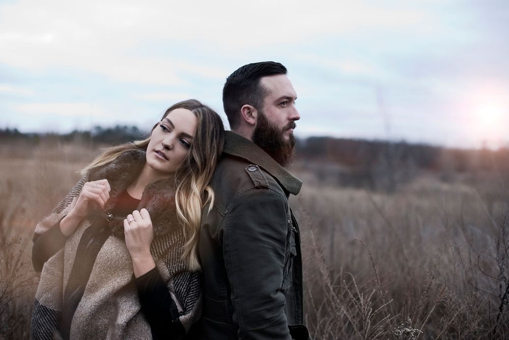 A couple stands back to back in a moody field at dusk with windswept hair and dramatic lighting.