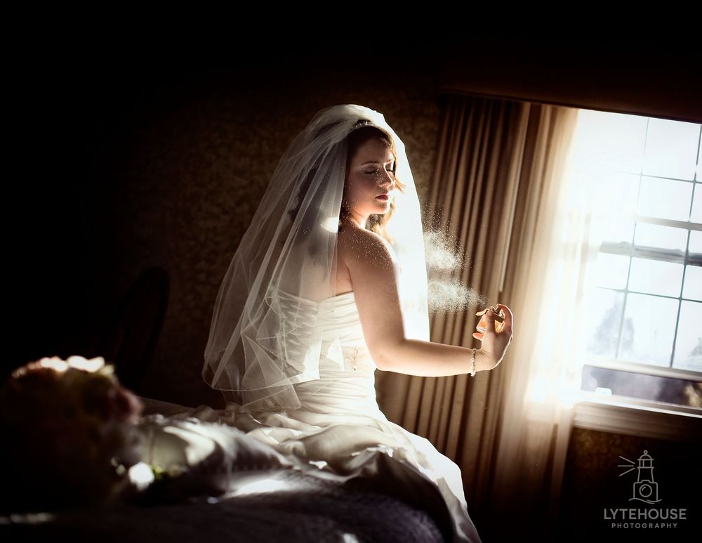 A bride sits near a sunlit window in a dark wood-paneled room wearing a white wedding dress and veil.