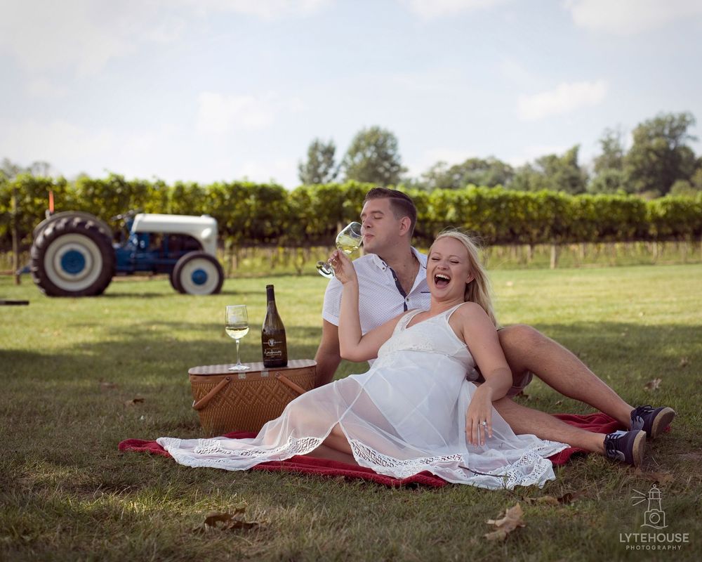 A couple in white outfits lounges on a blanket in front of a vineyard during a romantic sunset photo session.