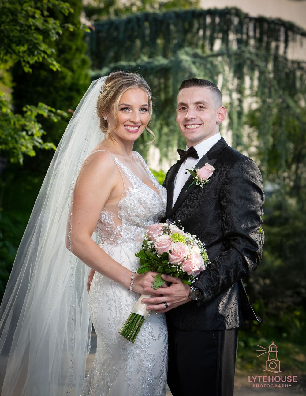 A bride and groom pose outdoors on their wedding day with the bride holding a pink floral bouquet.