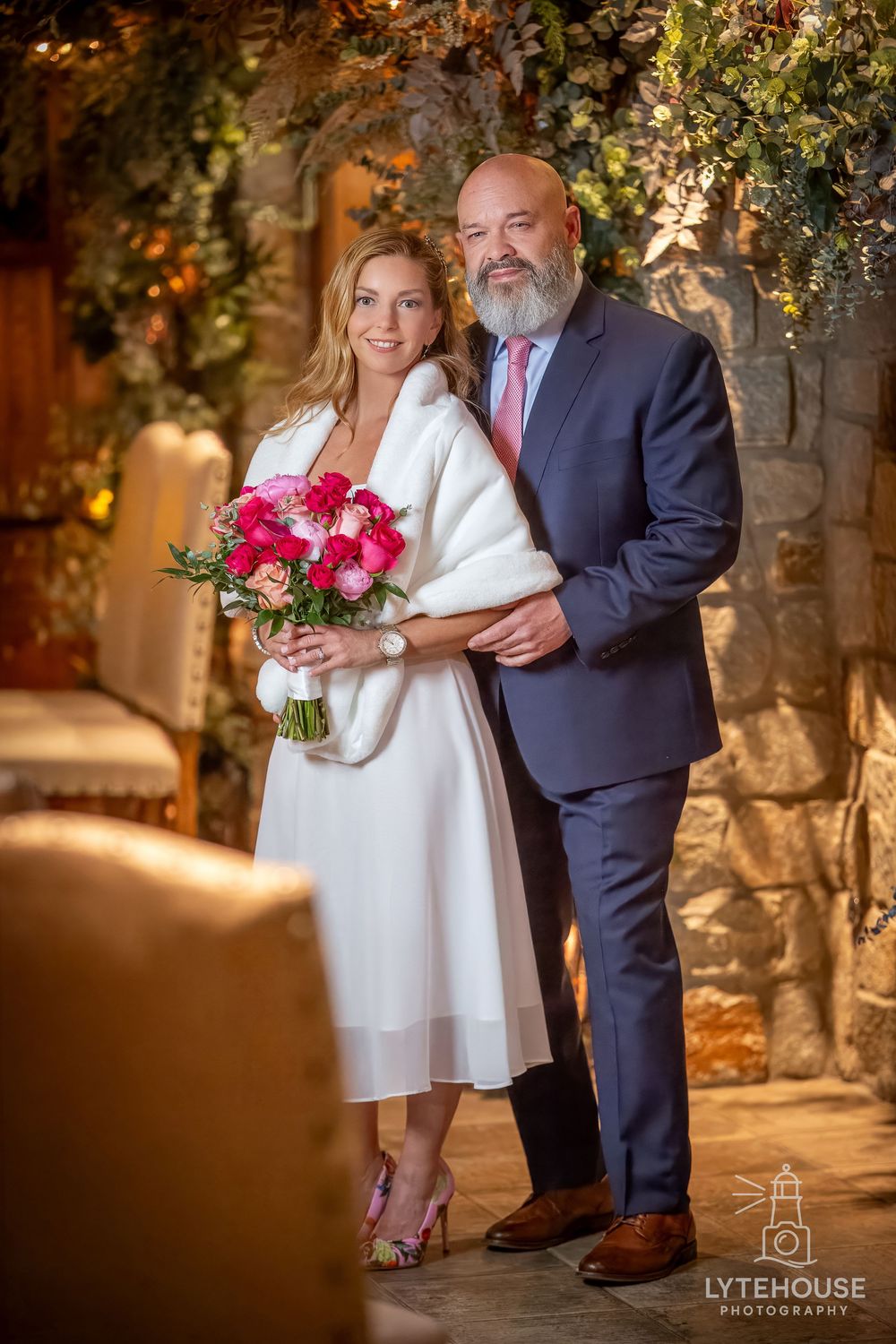 A couple poses together at a wedding venue with stone walls, floral decor, and the bride holding a vibrant pink bouquet.