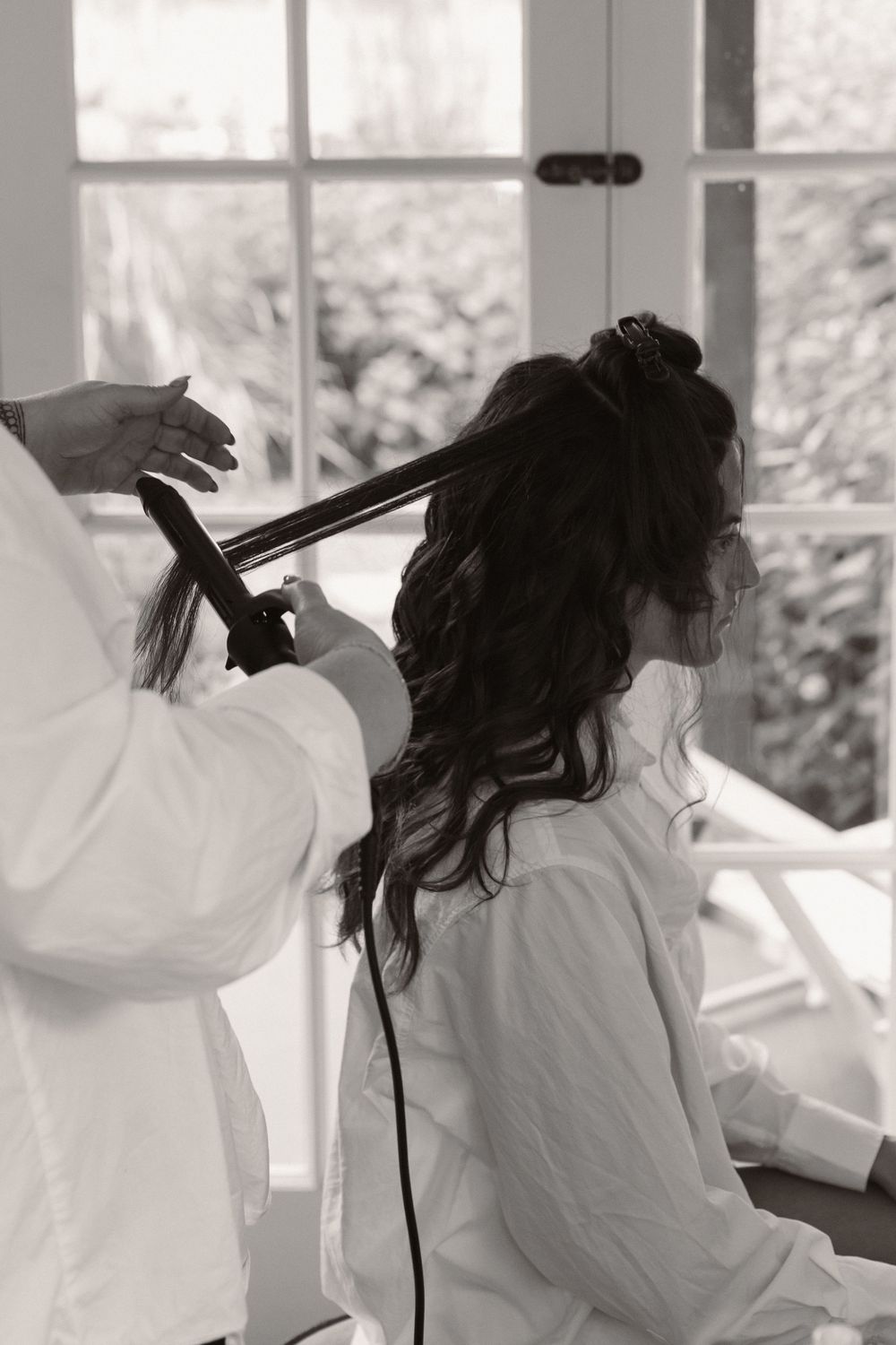 Hair stylist using flat iron to style long dark wavy hair in front of bright window.