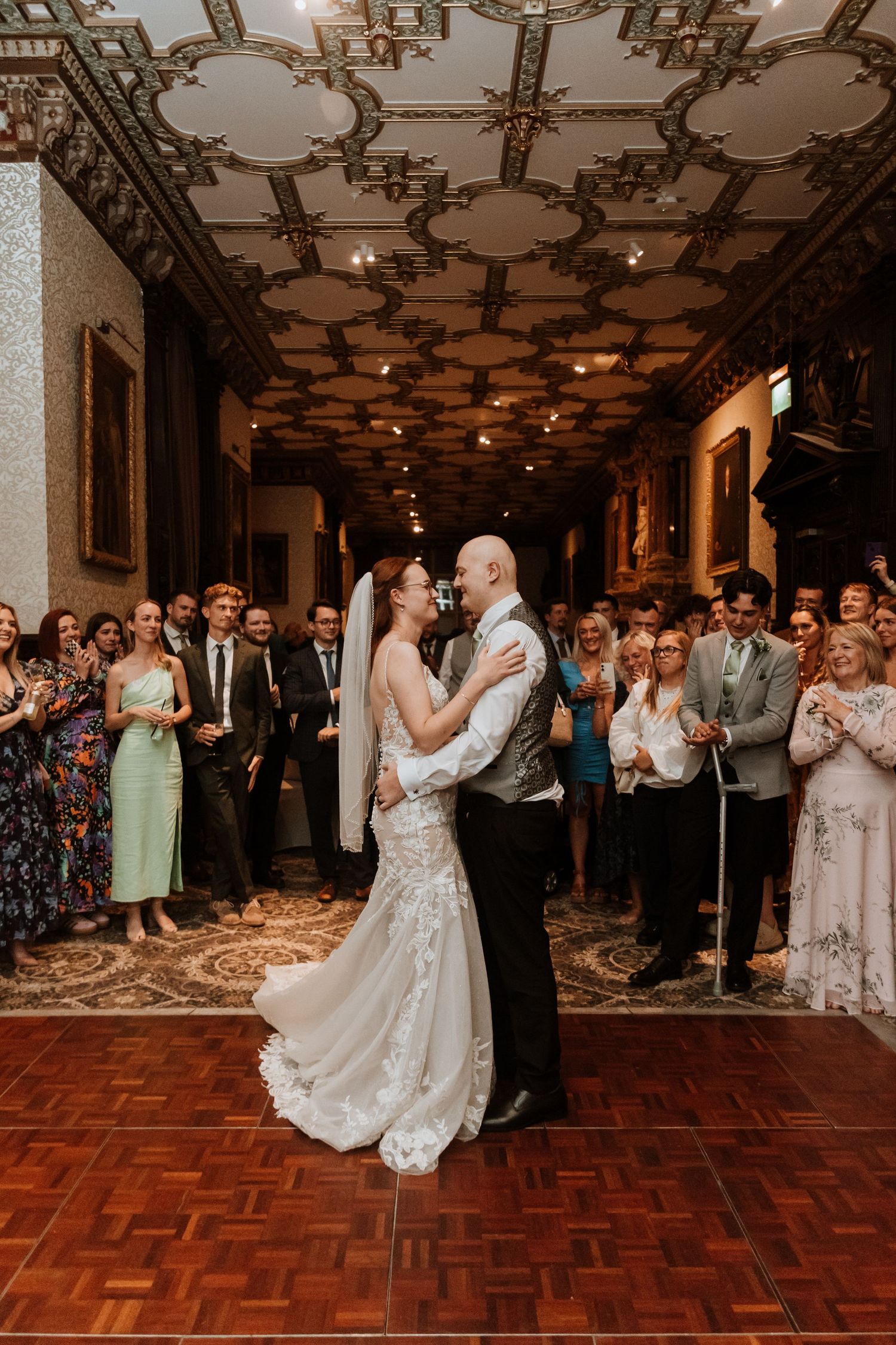 Newlyweds share their first dance on ornate parquet floor beneath elegant carved ceiling as wedding guests look on at Crewe Hall