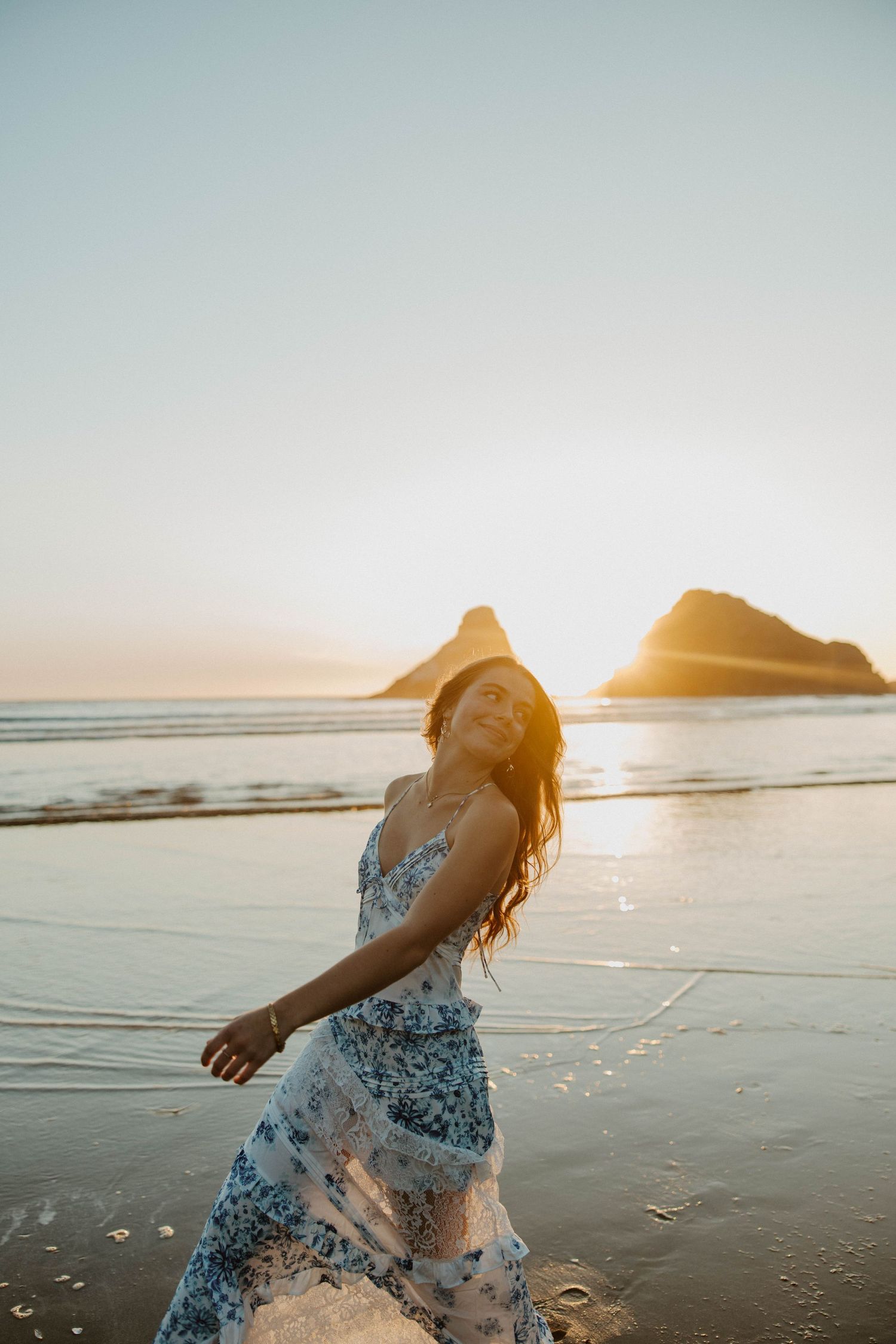 A floral dress twirls on a beach at golden hour with islands silhouetted in the background.