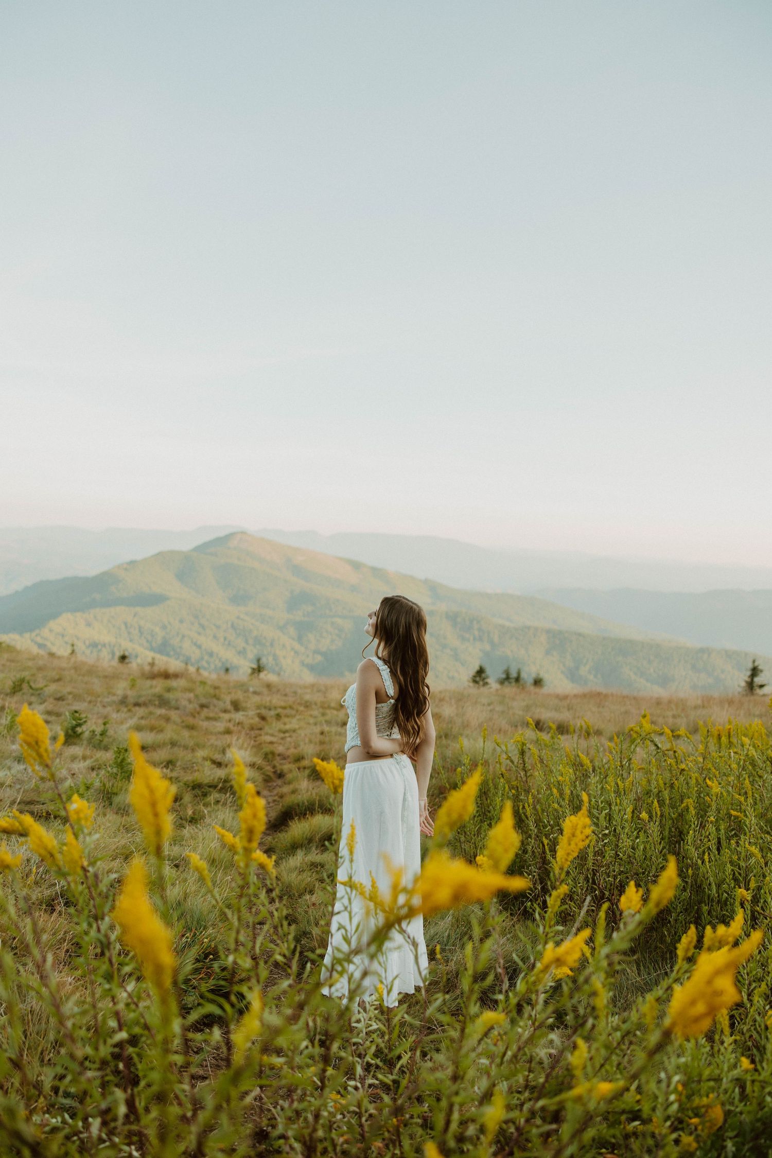 White dress flowing in mountain meadow surrounded by yellow wildflowers.