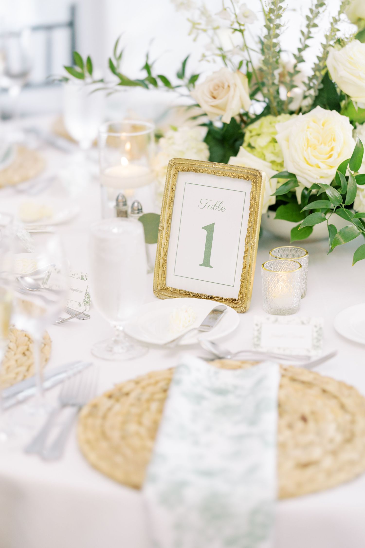 Elegant wedding table setting with gold framed table number one, white roses, and rattan charger plates.