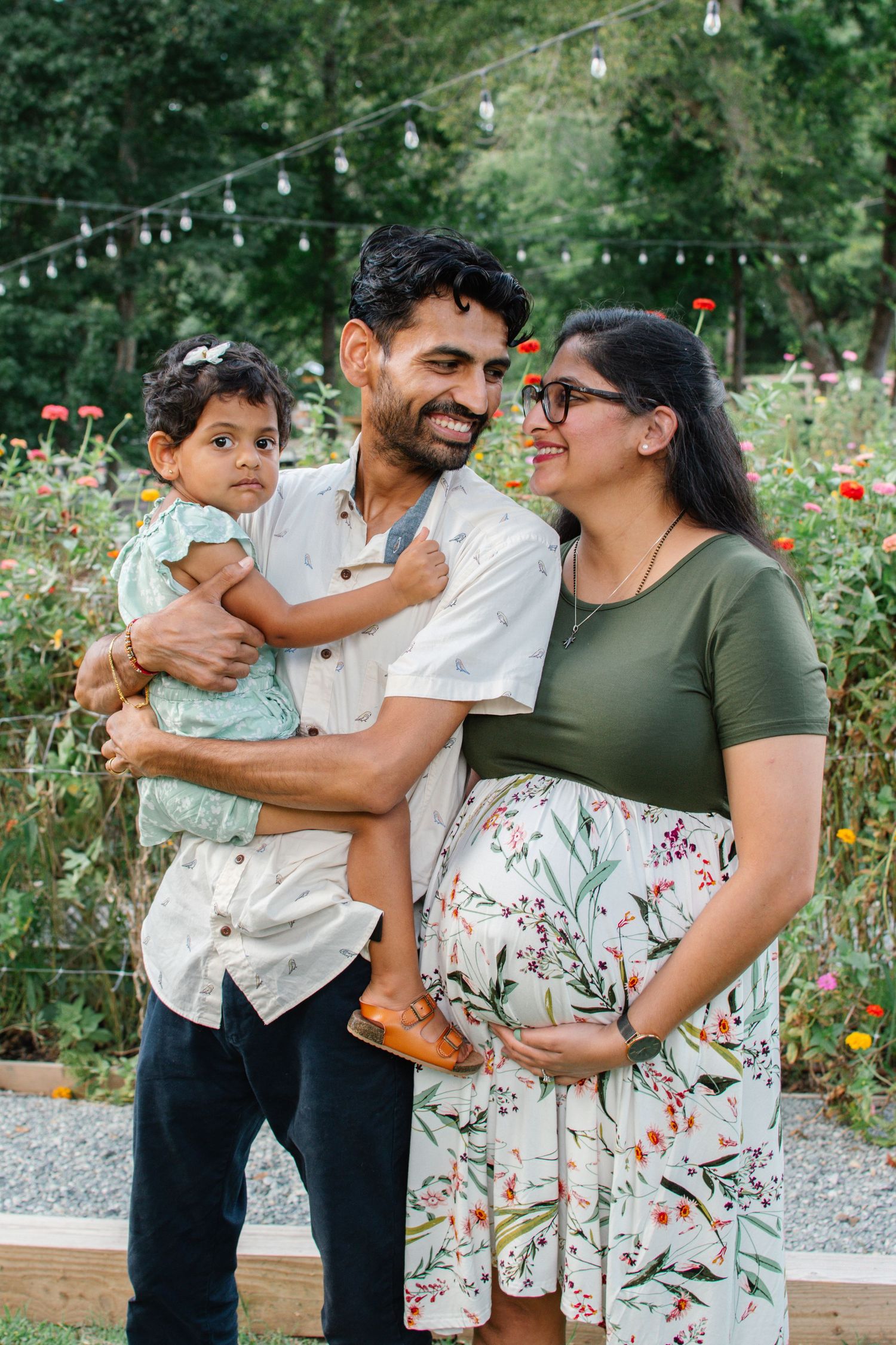 A smiling pregnant South Asian woman in floral skirt poses with her husband holding their toddler in a garden.