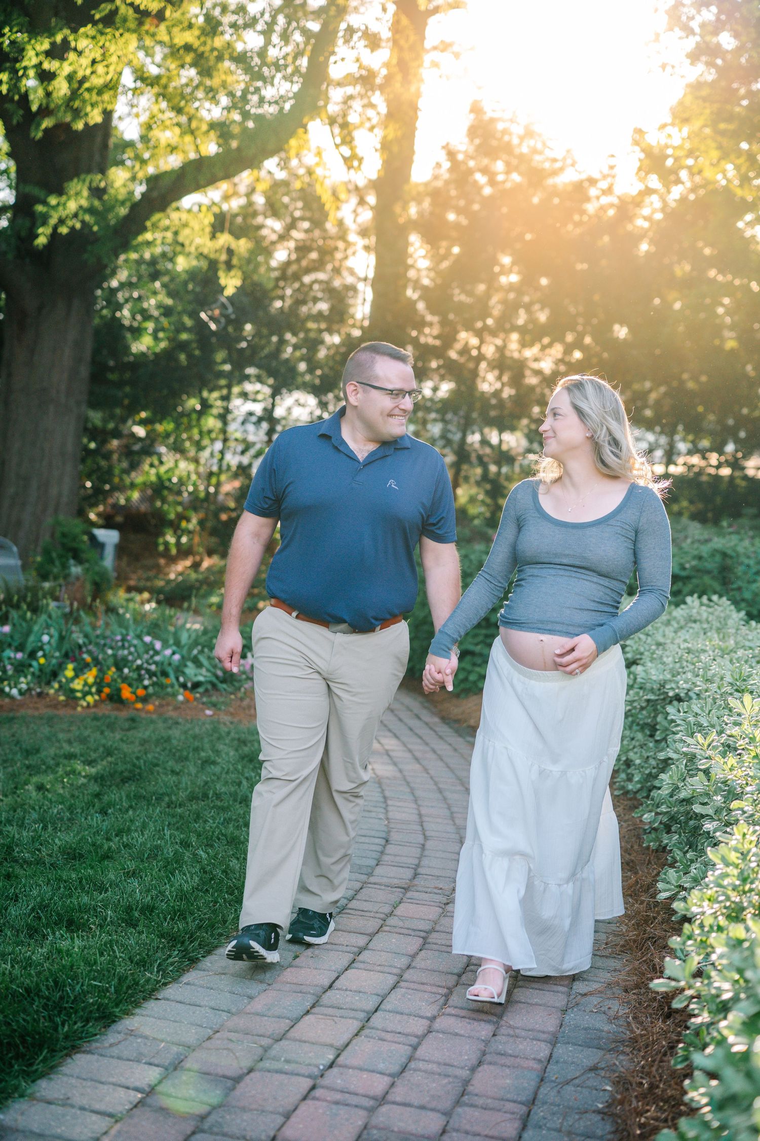 A happy expectant couple walks hand-in-hand along a brick garden path in warm golden sunlight.