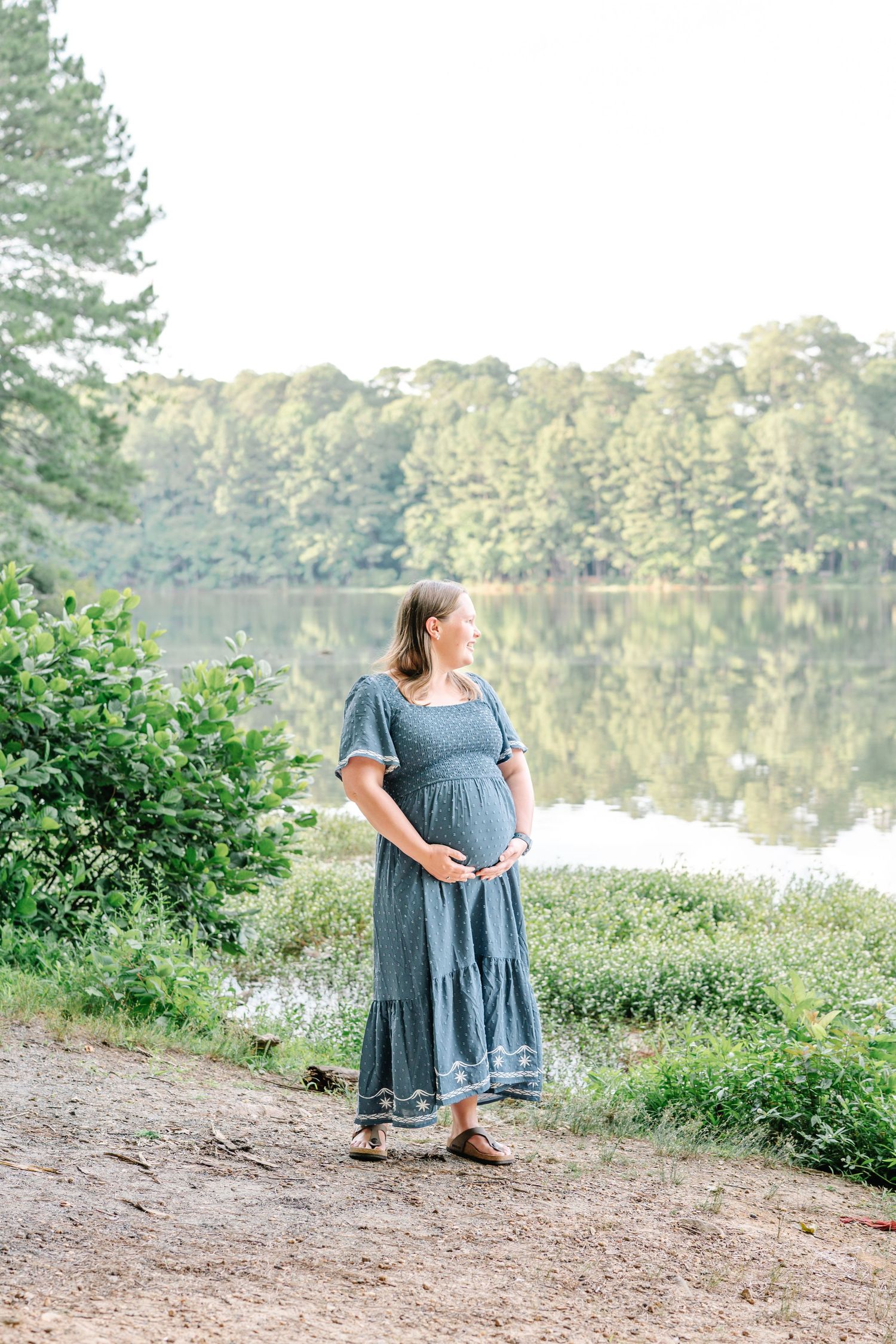 Pregnant woman in blue dress cradling baby bump by a serene lake surrounded by lush green trees.
