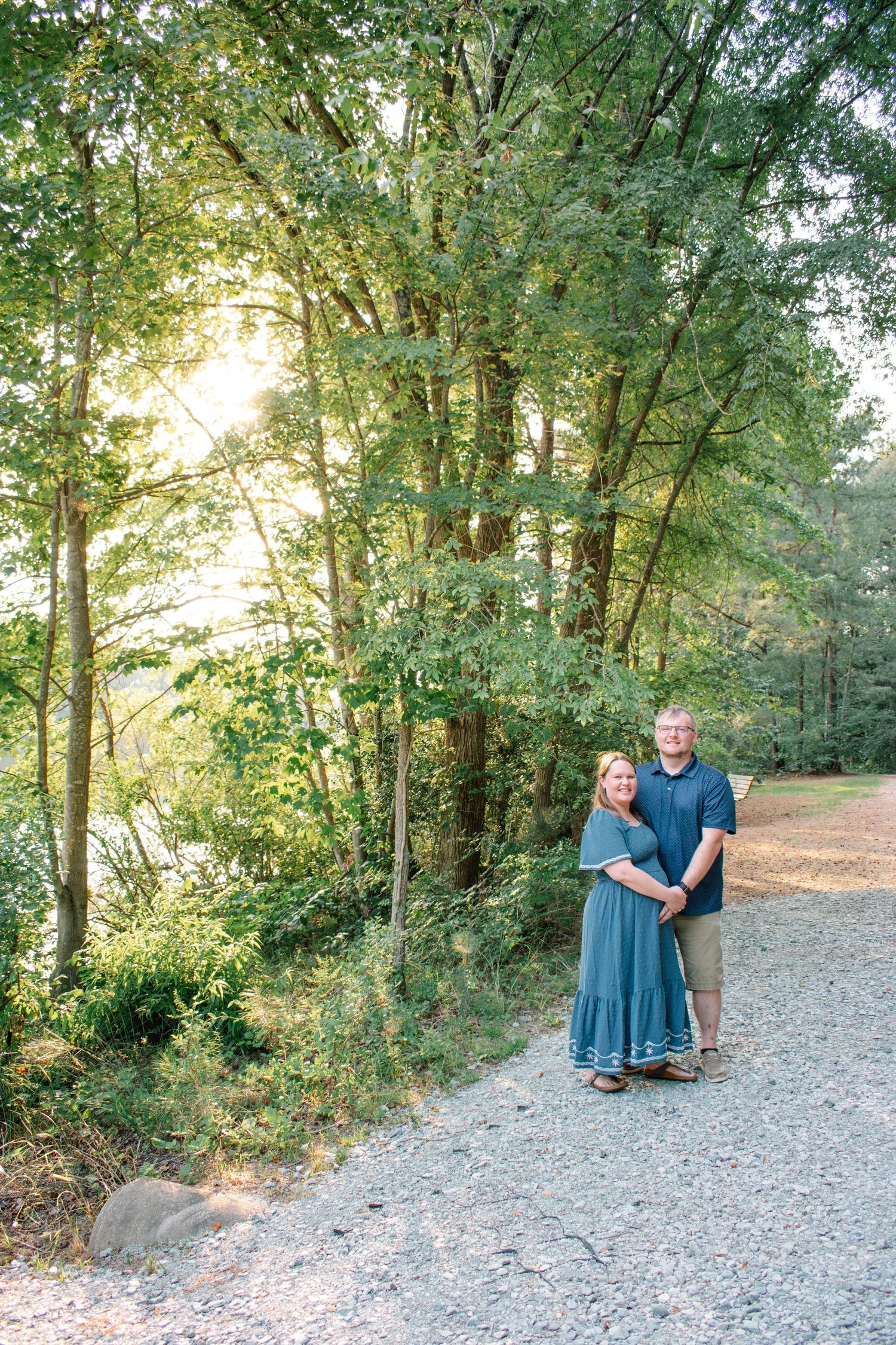 A couple smiles together on a gravel path surrounded by lush green trees with warm sunlight filtering through.